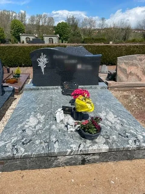 Monument funéraire en granit gris veiné avec plaques et fleurs dans un cimetière.